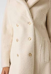 Roomkleurige faux bontjas met een double-breasted ontwerp, voorzien van goudkleurige knopen, ingesneden revers en zijzakken. Zachte, gestructureerde stof.