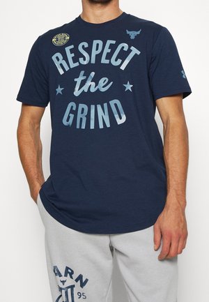 Mann trägt ein marineblaues T-Shirt mit dem Schriftzug „Respect the Grind“ und Logos, kombiniert mit hellgrauen Jogginghosen mit grafischen Texten und Symbolen.
