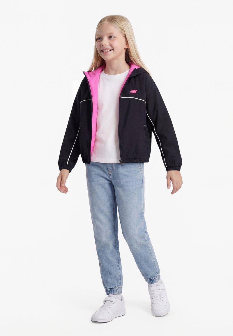 Χαμογελαστή νεαρή κοπέλα που φοράει μαύρο και ροζ μπουφάν, λευκό μπλουζάκι, ανοιχτό μπλε τζιν και λευκά αθλητικά παπούτσια, στέκεται σε λευκό φόντο.