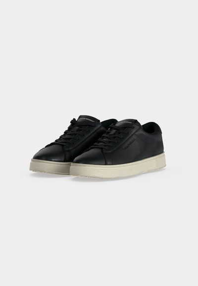 Paire de baskets basses en cuir noir avec lacets noirs et semelles épaisses en caoutchouc blanc sur fond clair.