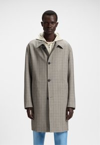 Cappotto a scacchi marrone con colletto, chiusura monopetto davanti e tre bottoni. Indossato sopra un hoodie chiaro, abbinato a jeans blu.