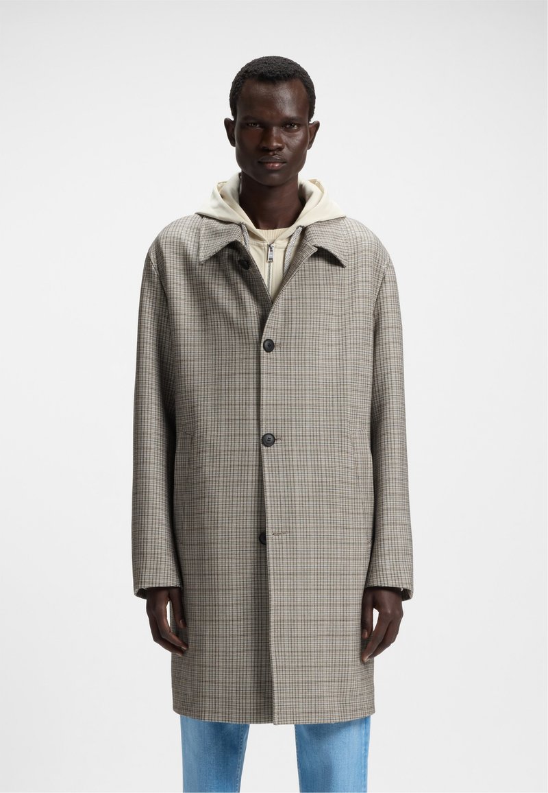Cappotto a scacchi marrone con colletto, chiusura monopetto davanti e tre bottoni. Indossato sopra un hoodie chiaro, abbinato a jeans blu.