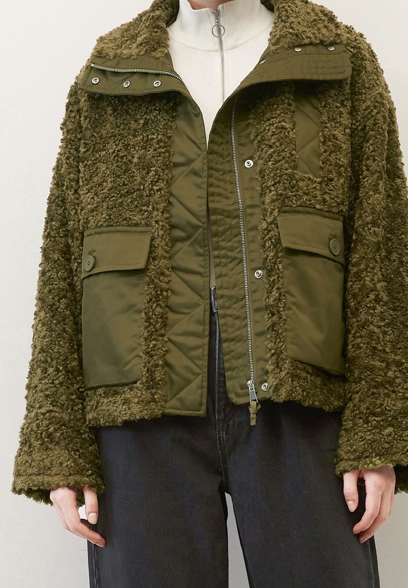 Oliven grøn jakke med en tekstureret, fluffy overflade og quiltet indre. Har trykknapper og påsyede lommer foran.