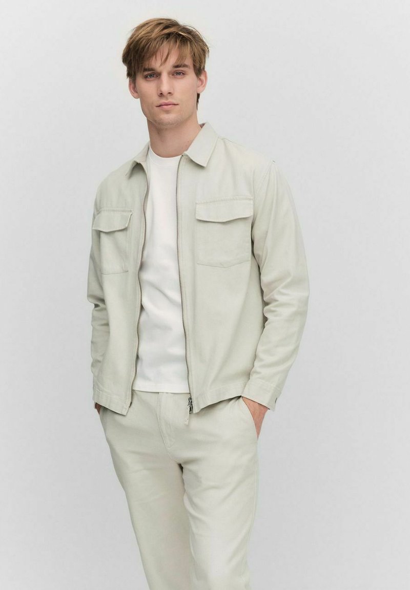 Lichtgrijze vest met rits en twee borstzakken, gecombineerd met een wit T-shirt en bijpassende lichtgrijze broek. Casual pasvorm, gladde textuur.