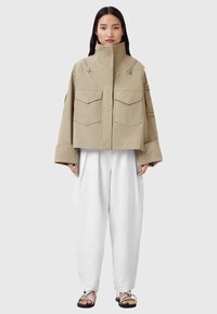Giacca cropped beige realizzata in materiale resistente, con colletto alto, due tasche sul petto e dettagli in zip argentata. Abbinata a pantaloni bianchi a gamba larga.