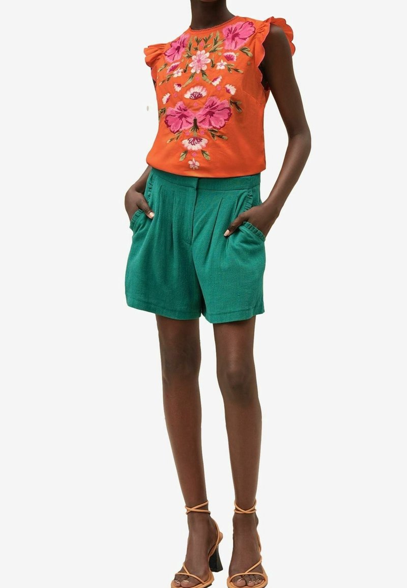 Femme portant un haut sans manches orange avec une broderie florale rose et un short plissé vert taille haute, mains dans les poches, debout avec des sandales.