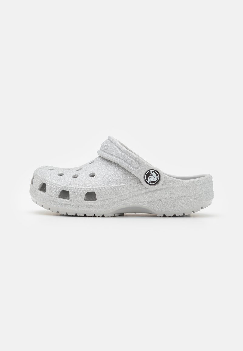 Des sabots Crocs blancs avec une surface texturée, des trous de ventilation et une lanière arrière ; présentant un accent de logo circulaire sur le côté.