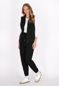 Gilet nero senza maniche con chiusura a zip, indossato sopra una camicia bianca a maniche lunghe, abbinato a pantaloni jogger neri e sneakers bianche.