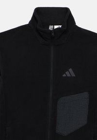 Μαύρο φλις μπουφάν με ψηλό γιακά, εμπρόσθιο φερμουάρ και λεπτομέρεια από καρό τσέπη. Διαθέτει διακριτικό λογότυπο στο στήθος.