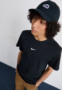 Crna pamučna majica s bijelim Nike logom na prsima, uparena s crnom kapom koja ima plavi i bijeli izvezeni logo.