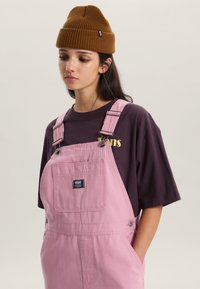 Roze denim tuinbroek met verstelbare bandjes en een voorzak, gecombineerd met een donkerpaarse t-shirt met een geel logo en een bruine gebreide muts.