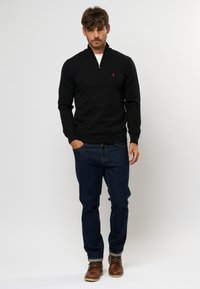 Schwarzer Pullover mit Reißverschluss und Rippbündchen, kombiniert mit dunkelblauen Jeans und braunen Lederstiefeln. Modell steht mit den Händen in den Taschen.