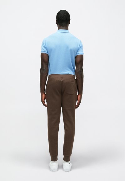 Καφέ φόρμες και ανοιχτό μπλε πουκάμισο πόλο από πίσω. Οι φόρμες διαθέτουν μια πίσω τσέπη. Άσπρα αθλητικά παπούτσια ολοκληρώνουν την εμφάνιση.