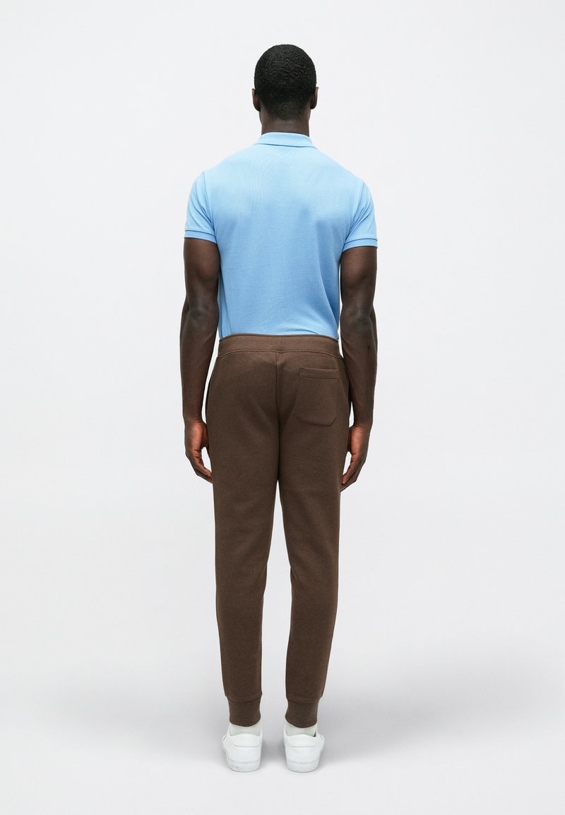 Braune Jogginghosen und ein hellblaues Poloshirt, von hinten gesehen. Die Jogginghosen verfügen über eine Gesäßtasche. WeißeSneaker vervollständigen das Outfit.