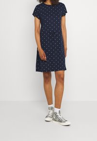 Robe bleu marine à manches courtes avec des pois blancs, taille cintrée et détail noué. Associée à des baskets montantes à motifs.