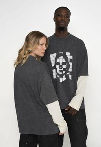 Para przerośniętych szarych t-shirtów z kontrastującymi białymi długimi rękawami. Jeden z nich ma graficzny nadruk twarzy, materiał o fakturze, luźny krój.