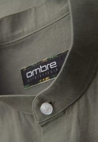 Camicia khaki con bottoni e colletto, con un bottone bianco e un'etichetta che recita "ombre quintessence" con un bordo decorato in verde.