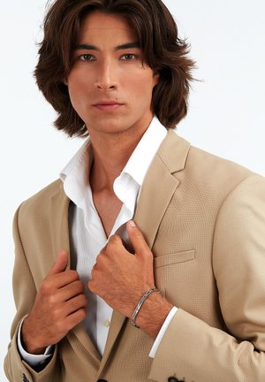 Beige kavaj över en vit skjorta, som visar en texturerad tyg. Ett silverarmband bärs på handleden, med händerna placerade vid kragen.