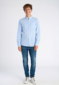 Lichtblauwe lange mouw knoopshirt met een borstzak, gecombineerd met middelblauwe jeans en witte sneakers met kleurrijke accenten.