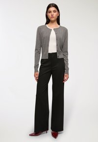 Cardigan grigio con applicazioni di perle, scollatura rotonda e chiusura a bottoni; abbinato a pantaloni a gamba larga neri a righe e tacchi bordeaux.