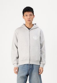 Un sweat à capuche gris zippé avec une poche avant, arborant un design graphique blanc sur le côté supérieur gauche et des poignets et un ourlet côtelés. Porté avec un jean bleu.