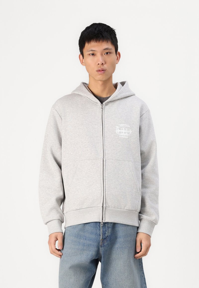 Un sweat à capuche gris zippé avec une poche avant, arborant un design graphique blanc sur le côté supérieur gauche et des poignets et un ourlet côtelés. Porté avec un jean bleu.