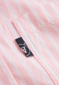 Țesătură cu dungi roz deschis și albă, cu un eticheta neagră ce prezintă un logo. Textura este netedă, detaliile includ cusături ordonate și un buzunar pe piept.