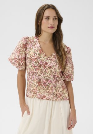 Žena u cvjetnoj ružičasto-bež bluzi s V-izrezom i puf rukavima stoji s jednom rukom u džepu krem suknje.