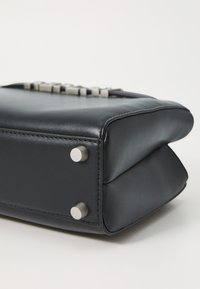 Sac en cuir noir de forme rectangulaire, avec un fond plat, des coins arrondis et des rivets en argent en bas pour la stabilité. Coutures détaillées visibles.