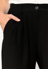 Pantaloni in lino neri con chiusura a bottone, tasche laterali e design plissettato. La texture appare morbida e leggera.