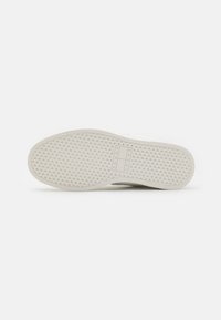 Fehér sneaker talp gyémántmintázatú barázdával és téglalap alakú "Tommy Jeans" logóval, amely a boltozatos rész közelében van megnyomva.
