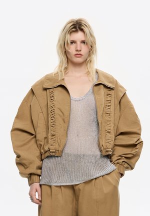 Modella con capelli biondi indossa una giacca oversize beige e un top in rete argentata, in piedi contro uno sfondo bianco con un'espressione seria.