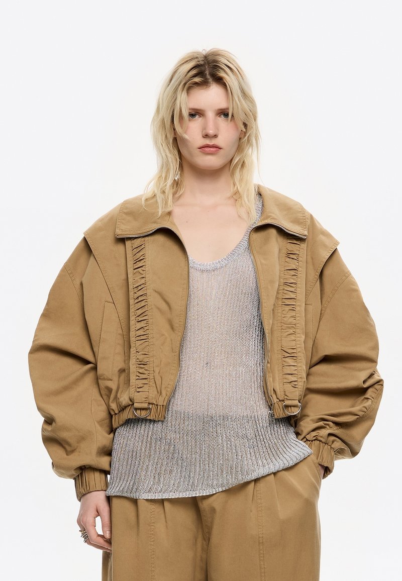 Modella con capelli biondi indossa una giacca oversize beige e un top in rete argentata, in piedi contro uno sfondo bianco con un'espressione seria.