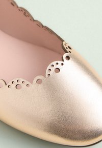 Chaussure plate en cuir doré avec un bord festonné, des découpes et une texture lisse et brillante. L'intérieur est rose clair.