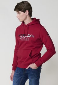 Felpa rossa con tasca a marsupio, cappuccio con cordino e testo stampato in bianco e nero. Indossata con jeans in denim blu, dal taglio slim.
