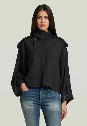 Vrouw met lang donker haar die een loszittende zwarte top met knopen en blauwe jeans draagt, staand met één hand in de zak tegen een effen achtergrond.