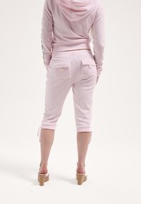 Világos rózsaszín, pamut keverék kapucnis pulóver húzózsinórral és zöld betűkkel. Hozzá illő, rövidnadrágszerű, két hátsó zsebes és megkötős részletekkel ellátott nadrág.