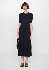 Abito a maglia a coste blu navy con maniche corte, corpo aderente, orlo svasato, indossato con una borsa verde scuro strutturata e scarpe Mary Jane marroni.