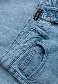 Jeans de mezclilla azul claro con cierre de botón metálico, detalles de costura y una pequeña etiqueta de marca negra en la cinturilla.