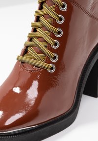 Botte en cuir verni marron brillant avec des lacets rayés jaunes et noirs, des œillets argentés, un bout effilé et un talon épais noir.