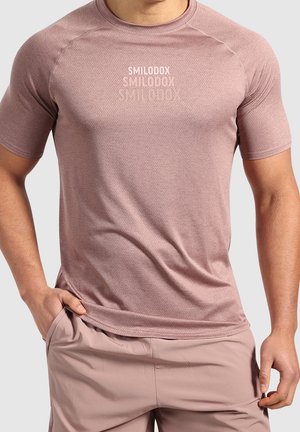 Mann trägt eng anliegendes, staubrosafarbenes, strukturiertes T-Shirt mit "SMILODOX"-Logo und passende hellrosa Shorts, steht mit den Händen nahe den Taschen.