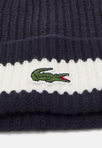 Kötött sapka bordás sötétkék anyagból, fehér vízszintes csíkkal és zöld krokodil logóval hímzéssel az elején.