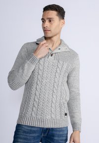 Pulover gri tricotat cu guler tip șal, butoni din lemn, manșete și tiv cu aspect rasucit. Purtați-l cu blugi albaștri, punând în evidență textura și detaliile de design.