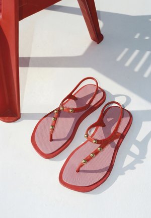 Sandales rouges et roses avec des clous dorés placées sur une surface blanche à côté des pieds rouges en plastique d'une chaise qui projettent des ombres.