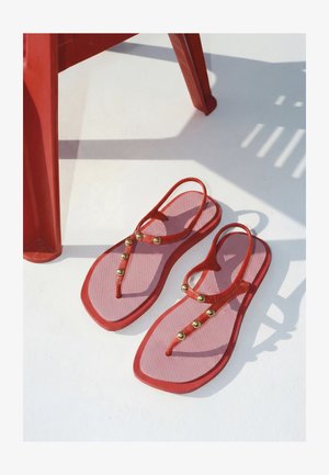 Rote und pinke Sandalen mit goldenen Nieten auf weißer Oberfläche neben roten Plastikstuhlbeinen, die Schatten werfen.