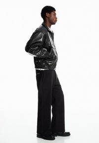 Zwarte leren jas met een getextureerde afwerking, gecombineerd met wijde zwarte broek en zwarte schoenen. Het model staat op de zij tegen een witte achtergrond.