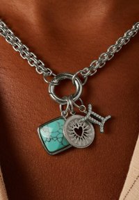 Zilveren ketting met een turquoise stenen bedel, een ronde metalen bedel met een hart, en een rhinestone sterrenbeeld symbool, allemaal met verschillende texturen.
