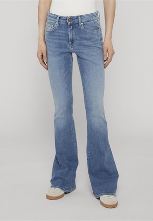 Donna con jeans a zampa di elefante azzurri chiari e sneakers bianche, in piedi davanti a uno sfondo semplice.