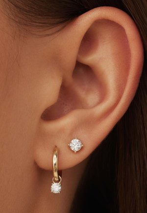 Gouden hoepeloorbel met een kleine hangende cubic zirconia, gecombineerd met een studoorbel met een grotere cubic zirconia, beide in een gouden tint.