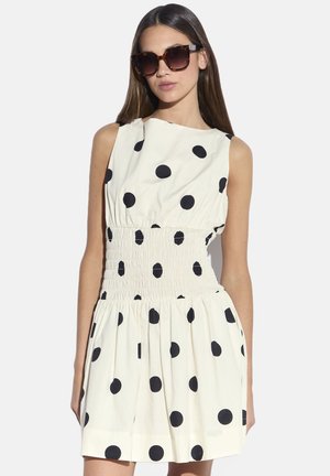 Abito a pois color crema con grandi puntini neri, caratterizzato da una vita arricciata e una gonna svasata. Modella con occhiali da sole oversize.